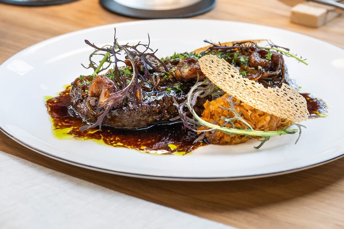 Teller mit kunstvoll angerichtetem Fleischgericht, daneben Schale mit Pommes und Glas Rotwein auf Holztisch
