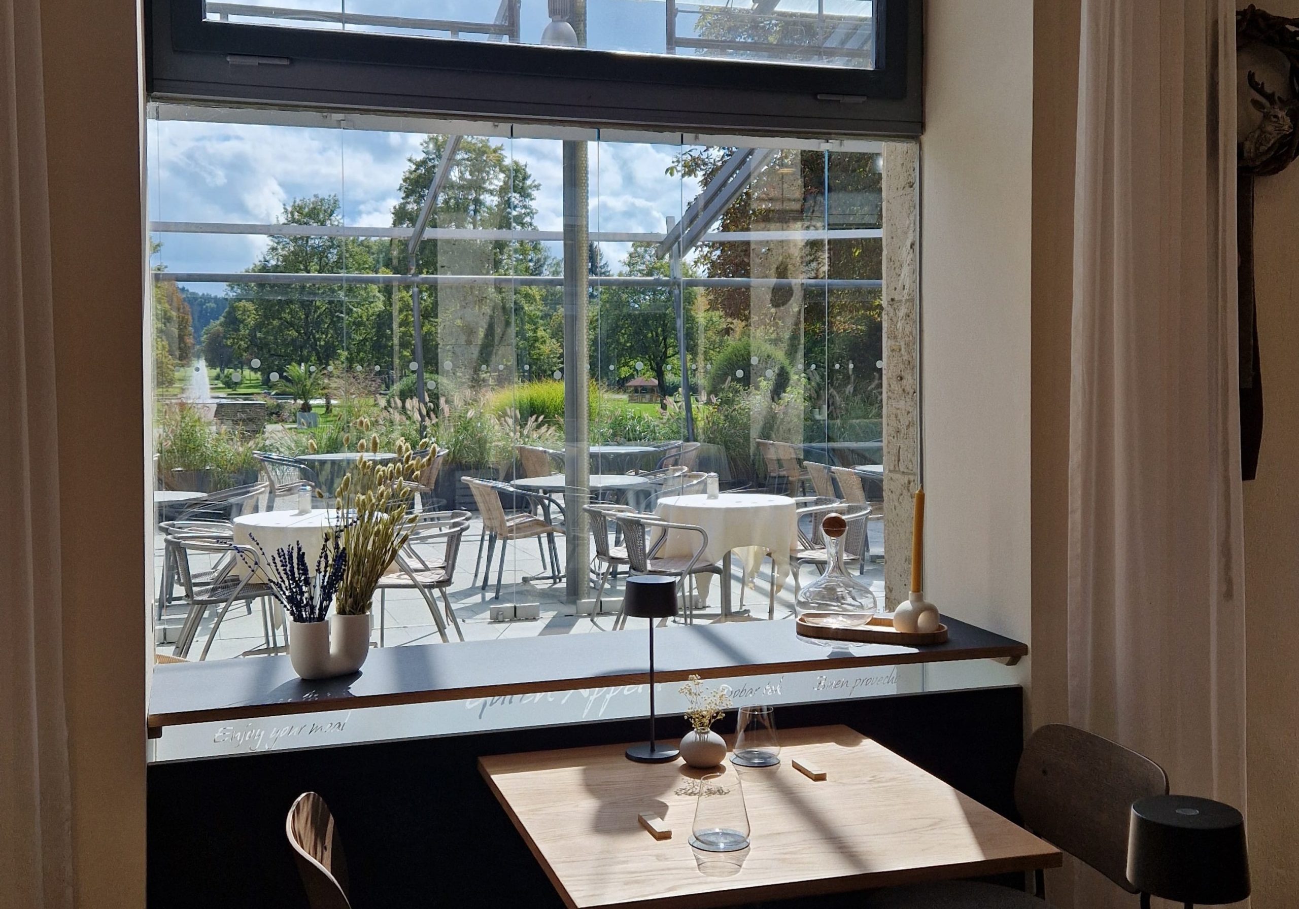 Blick durch ein großes Fenster auf eine Terrasse mit Tischen und Stühlen, im Vordergrund ein Tisch mit Glas, Vase und Kerze im Innenraum