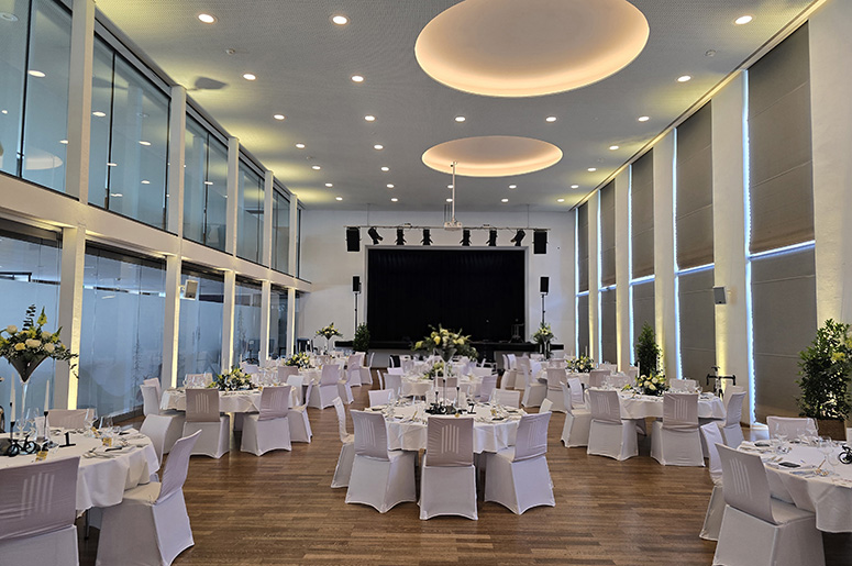 Großer Saal mit runden Tischen und weiß bezogenen Stühlen vor einer Bühne mit schwarzem Vorhang und großen runden Deckenleuchten